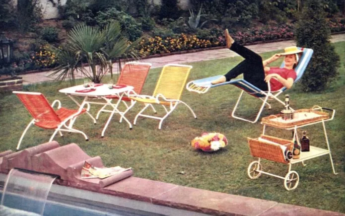 A stylish 1960s outdoor patio set by ILSE Möbel, featuring colorful woven chairs, a white metal table, and a woman lounging on a recliner in a lush garden.